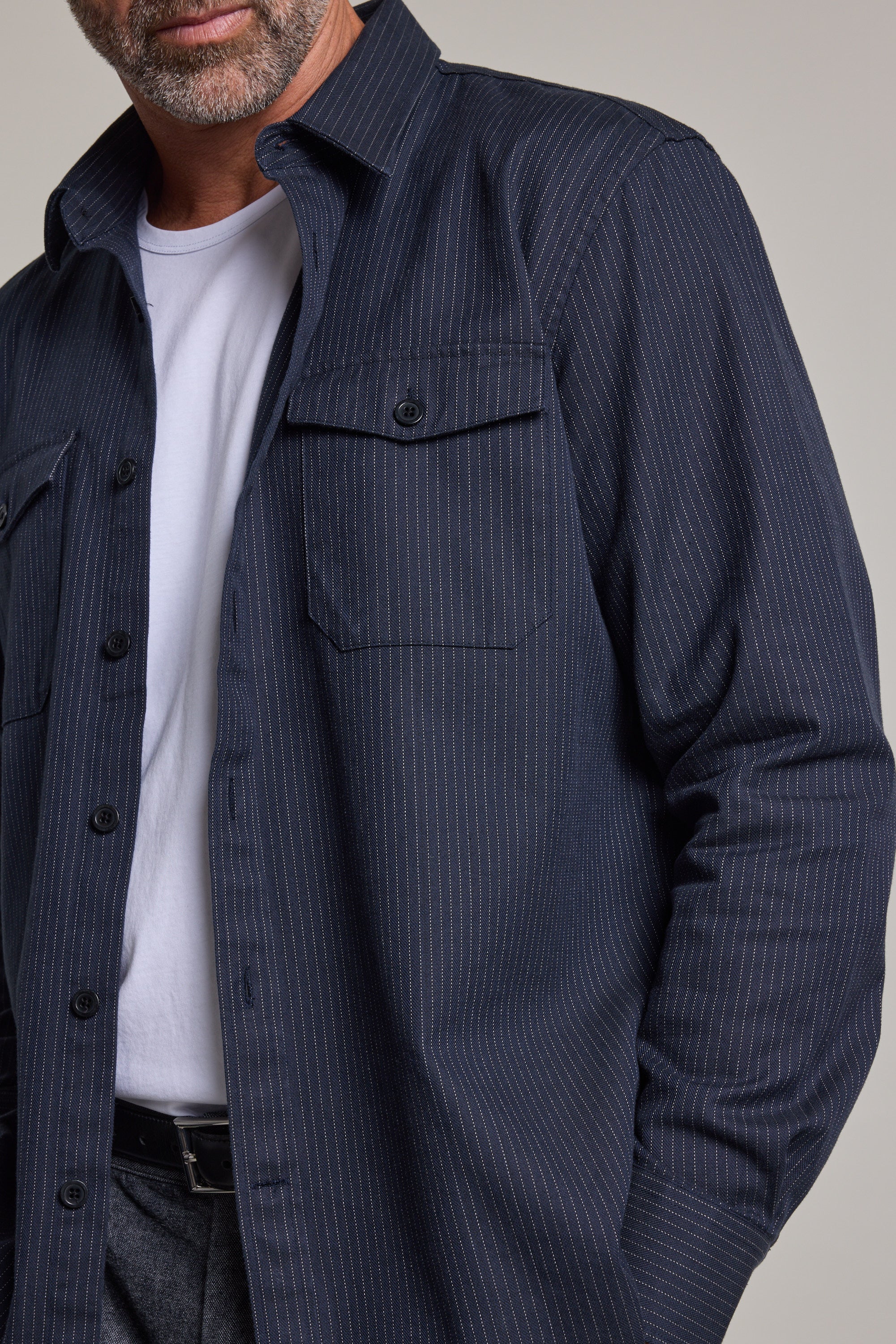 A man wears the Barkers Timothy Stripe Overshirt—a dark blue, cotton pinstripe shirt with chest pockets and black buttons—over a white t-shirt. Only his lower face and torso are shown.