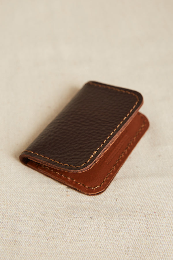The Parisian Leather Fold Wallet by Parisian, a small handmade leather wallet with visible stitching, rests on a beige fabric surface.