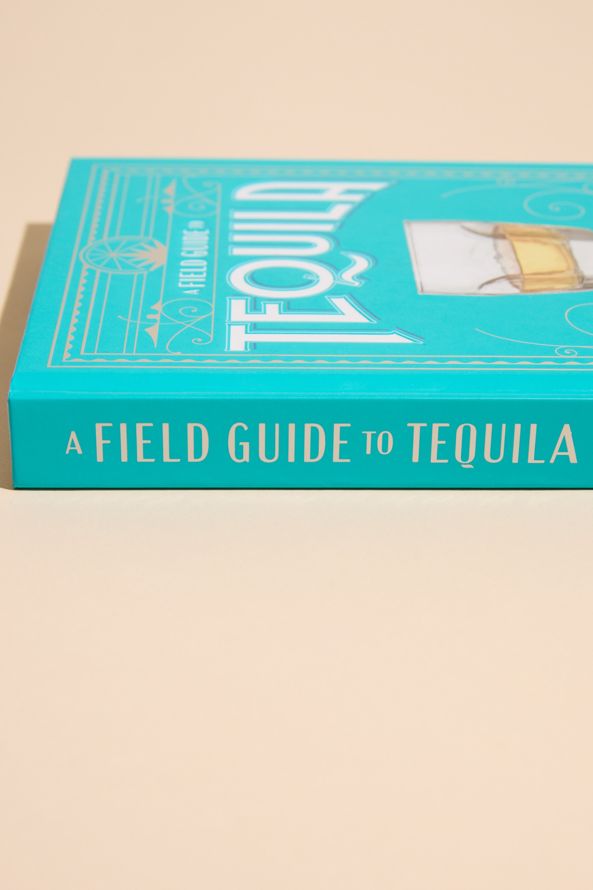 A Field Guide To Tequila by Other rests on a beige surface with its turquoise spine and partial cover visible—an ideal companion for exploring artisanal tequila.