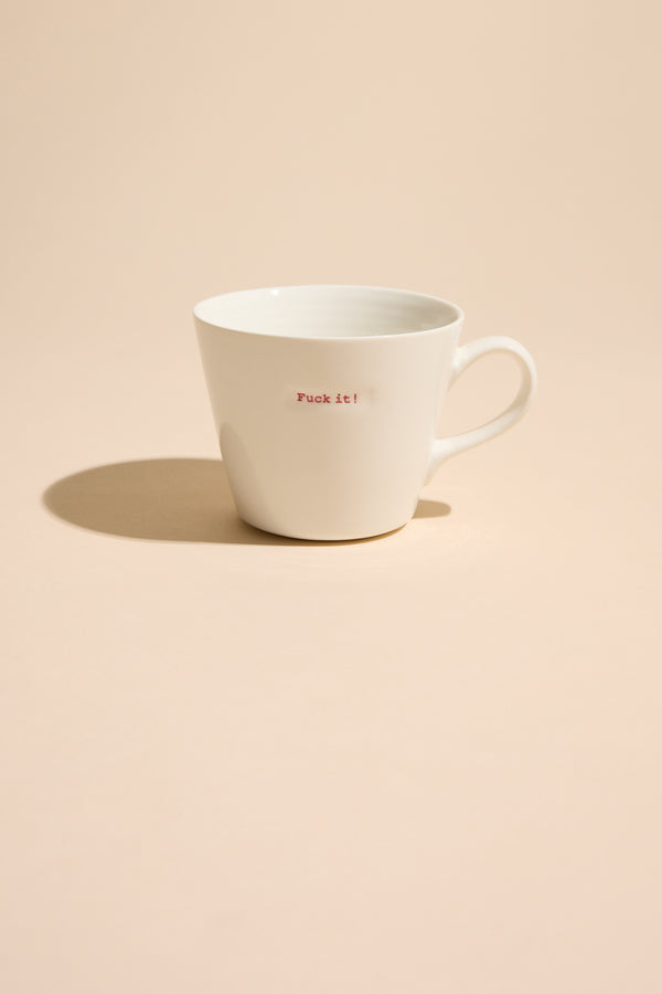 The Bucket Mug - F*ck it! by Other is a white ceramic mug with bold small red text, sitting on a beige surface and casting a shadow—a striking designer piece with attitude.