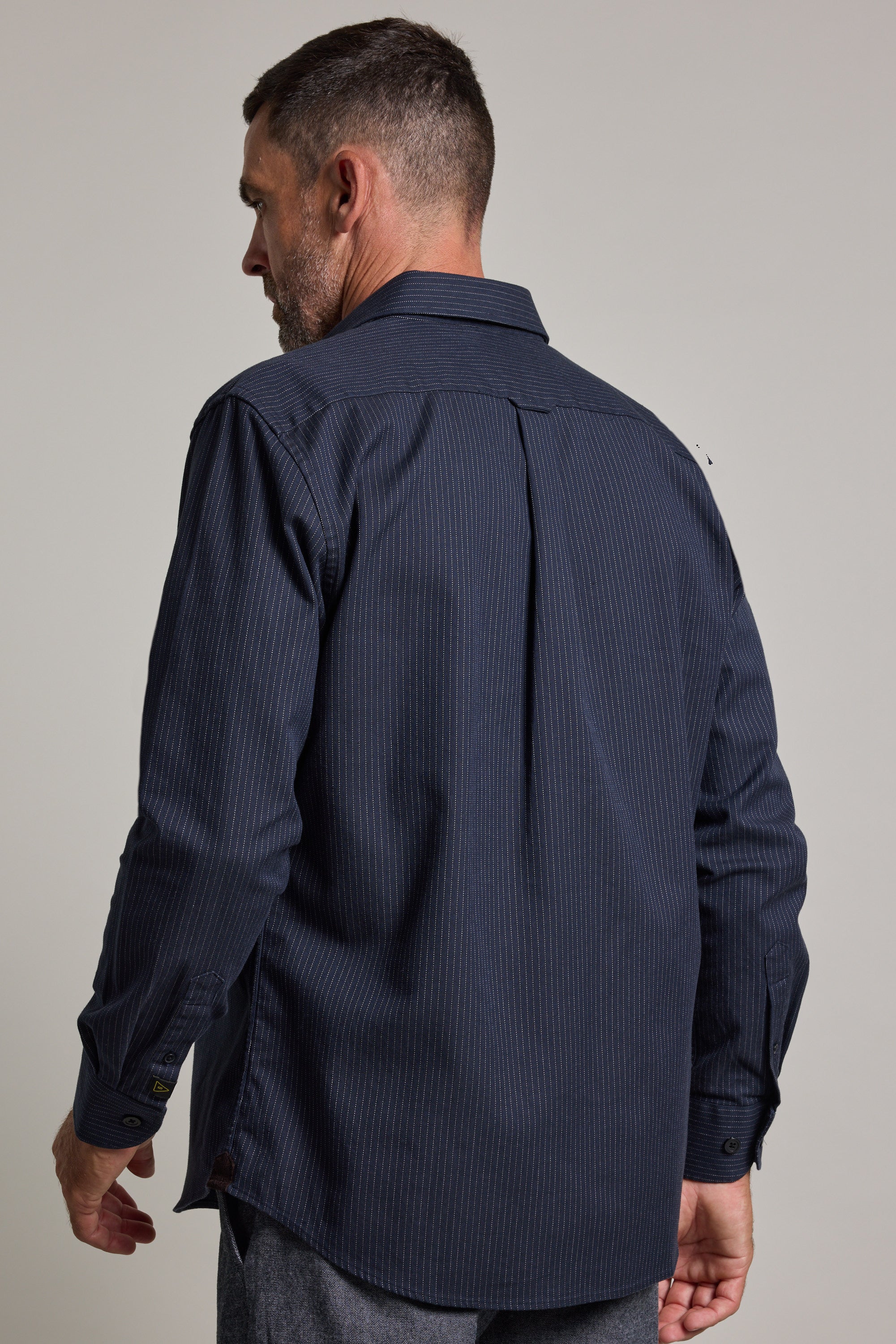 A man with short hair and a beard wears a dark blue Barkers Timothy Stripe Overshirt with a subtle pinstripe pattern. He is facing away from the camera and looking to his left.