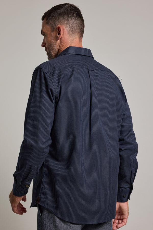 A man with short hair and a beard wears a dark blue Barkers Timothy Stripe Overshirt with a subtle pinstripe pattern. He is facing away from the camera and looking to his left.