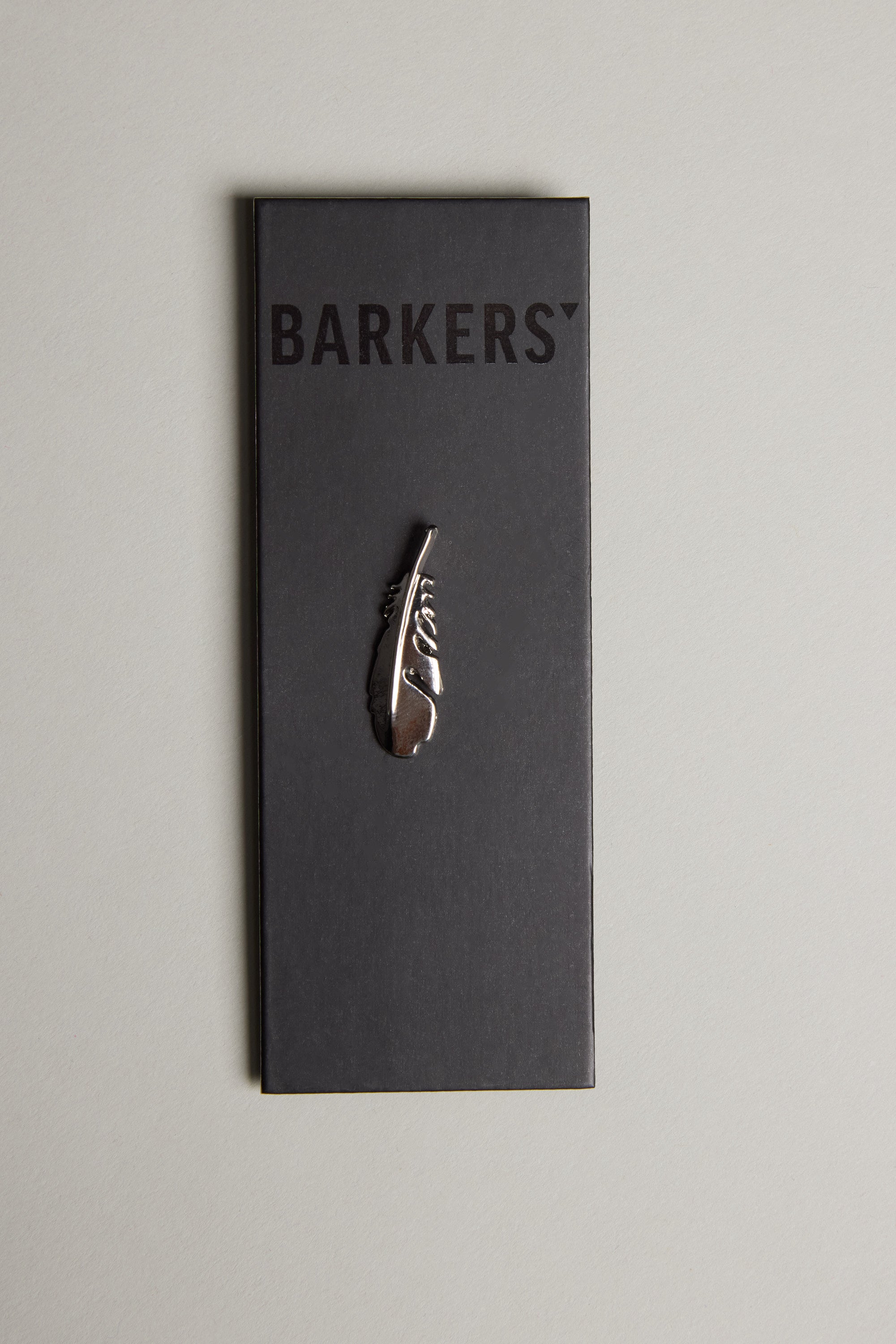 The Feather Touch Lapel Pin by Barkers features a polished silver tone feather engraved with “Story,” presented on a black card labeled Barkers and displayed on a light grey surface.