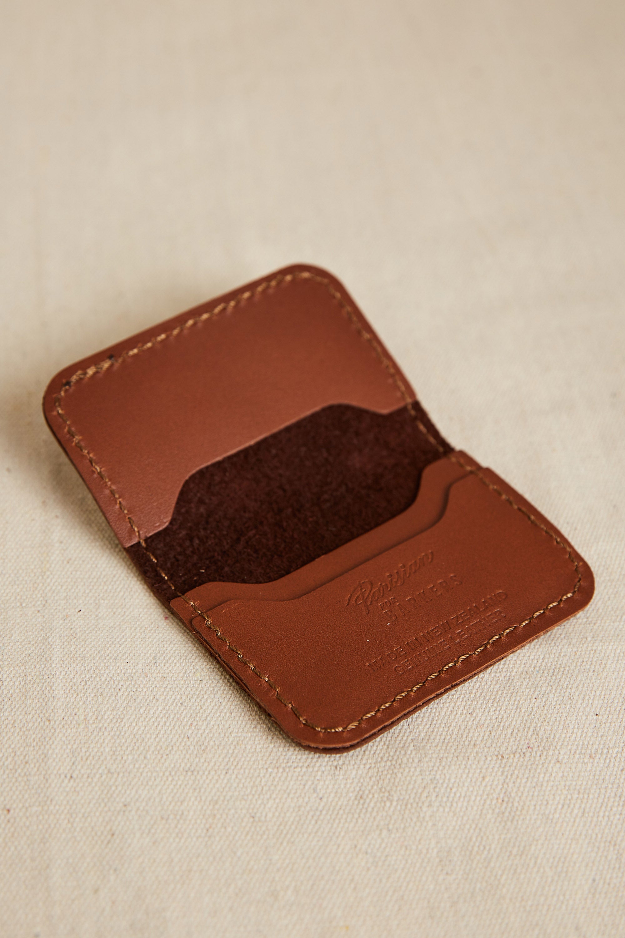 The Parisian Leather Fold Wallet by Parisian, crafted in brown New Zealand leather with stitched edges, is open to reveal two inner pockets and rests on a beige fabric surface.