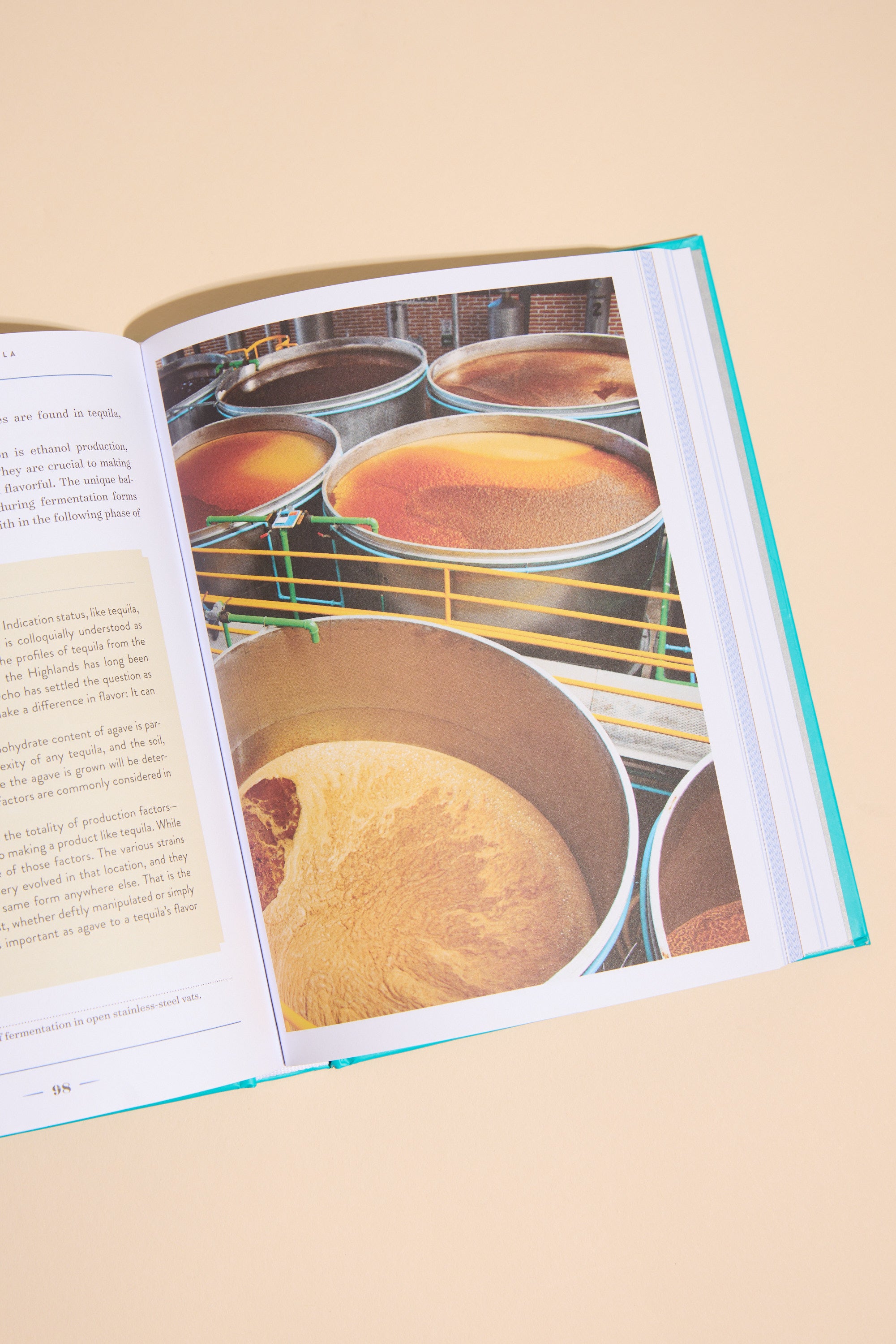 An open copy of "A Field Guide To Tequila" by Other shows a photo of industrial tanks with brownish liquid, likely related to tequila production, alongside printed text. The book is placed on a beige surface.