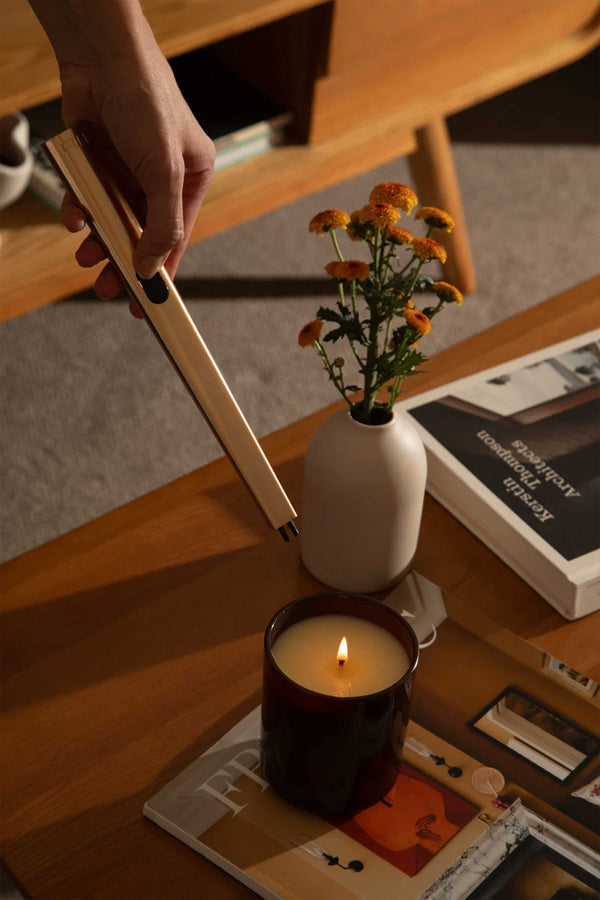 A hand holds the Flint Rechargeable Lighter 2.0 above a lit candle on a wooden table, next to a small vase with yellow flowers and several books and magazines.