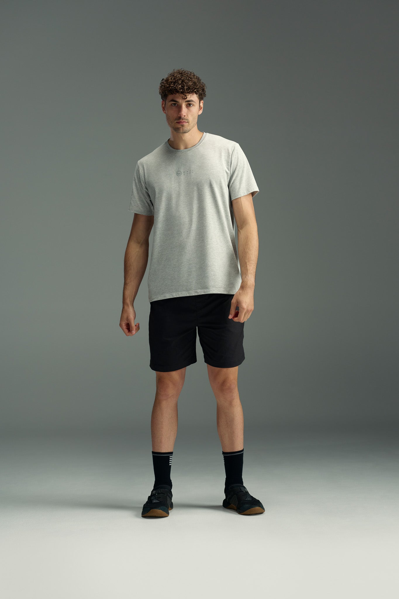 A man with curly hair stands against a gray background, wearing a light gray t-shirt and black WearEver Agile Short by WearEver—7-inch lightweight stretch shorts—paired with black socks and sneakers.
