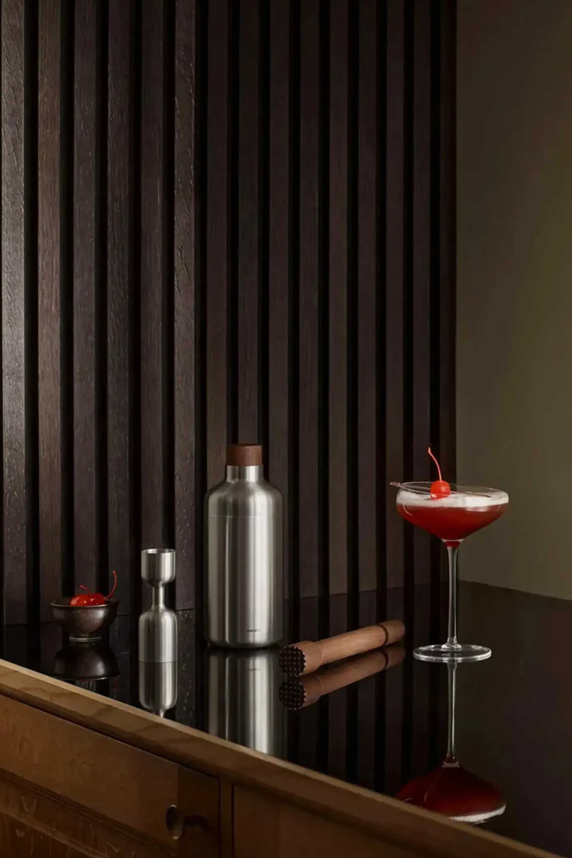 A stylish bar scene featuring the Liquid Lounge Cocktail Jigger by Other, a metal shaker, wooden muddler, bowl of cherries, and a cherry-garnished cocktail atop a glossy black counter framed by dark wood paneling.
