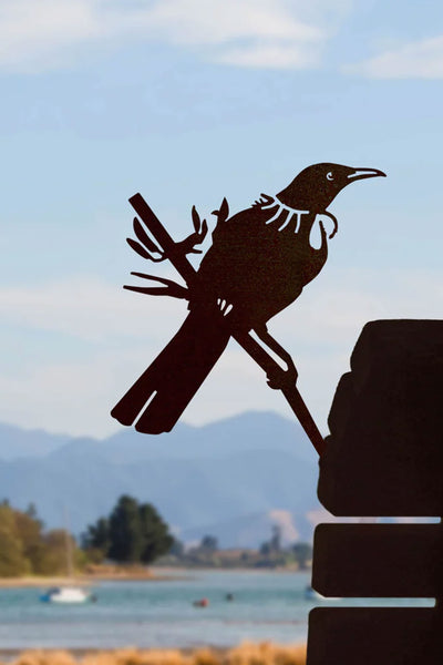 The Metal Bird Tui by Other features a Corten steel tui with a signature collar, perched on a branch against blurred mountains, trees, and blue sky—an elegant piece of New Zealand garden art.